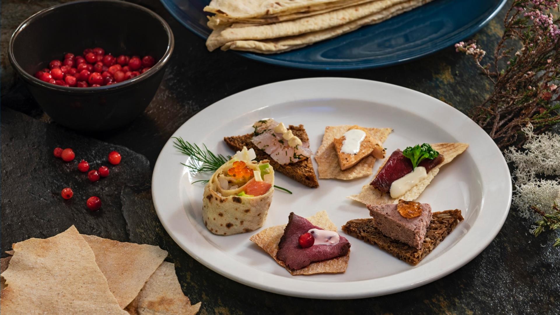 Assorted open sandwiches on a plate with herbs and berries, on a dark tabletop with flatbread nearby