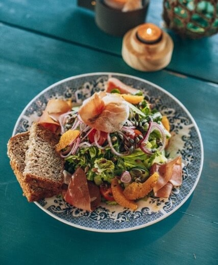 Plate of mixed salad with cured meat and toasted bread on a table, with a wooden salt grinder nearby