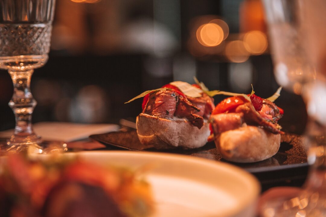 Grilled meat and bread rolls cooking on a restaurant barbecue grill, with wine glasses in the foreground.