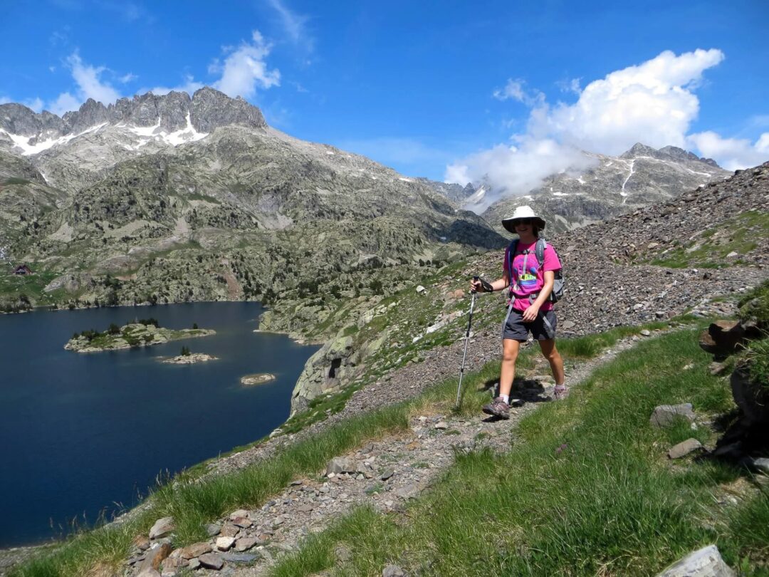 Woman hiking a mountain trail beside a lake under rocky peaks, holding trekking poles
