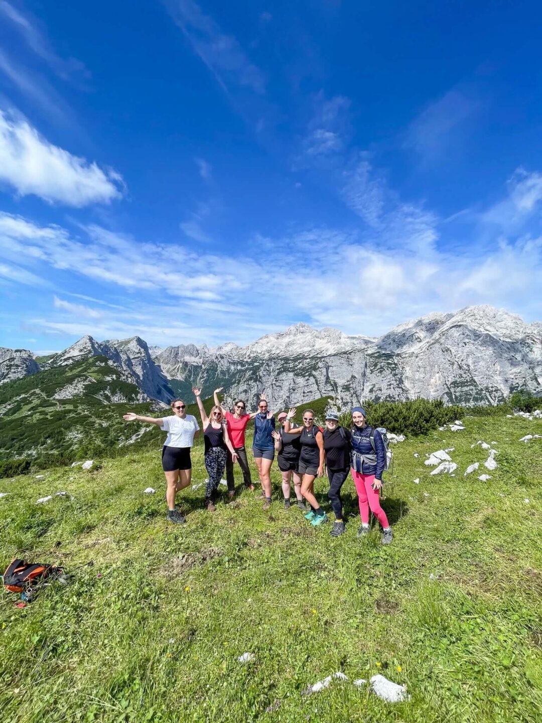 Women hiking on an alpine trail with backpacks in front of snowy mountains under a blue sky.