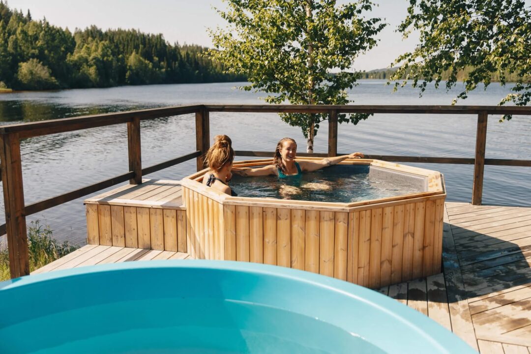 Two women relaxing in an outdoor hot tub by a lake in Lapland, surrounded by forest trees