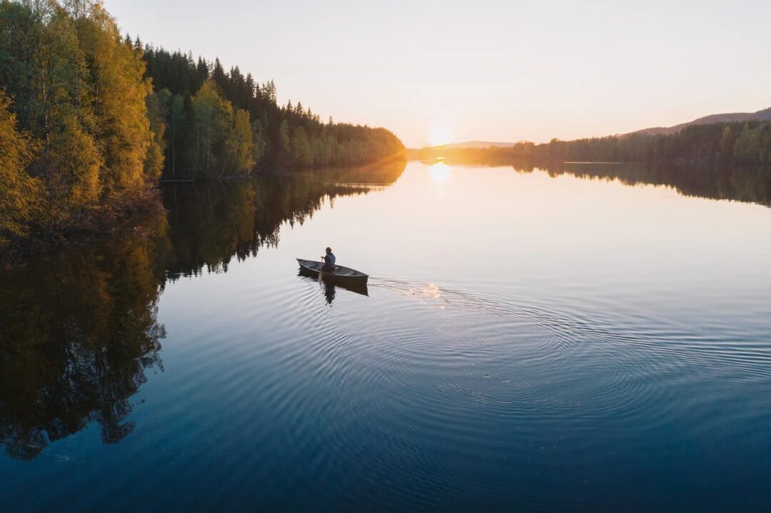 Canoe on calm lake at sunrise, with forest-lined shores and misty hills