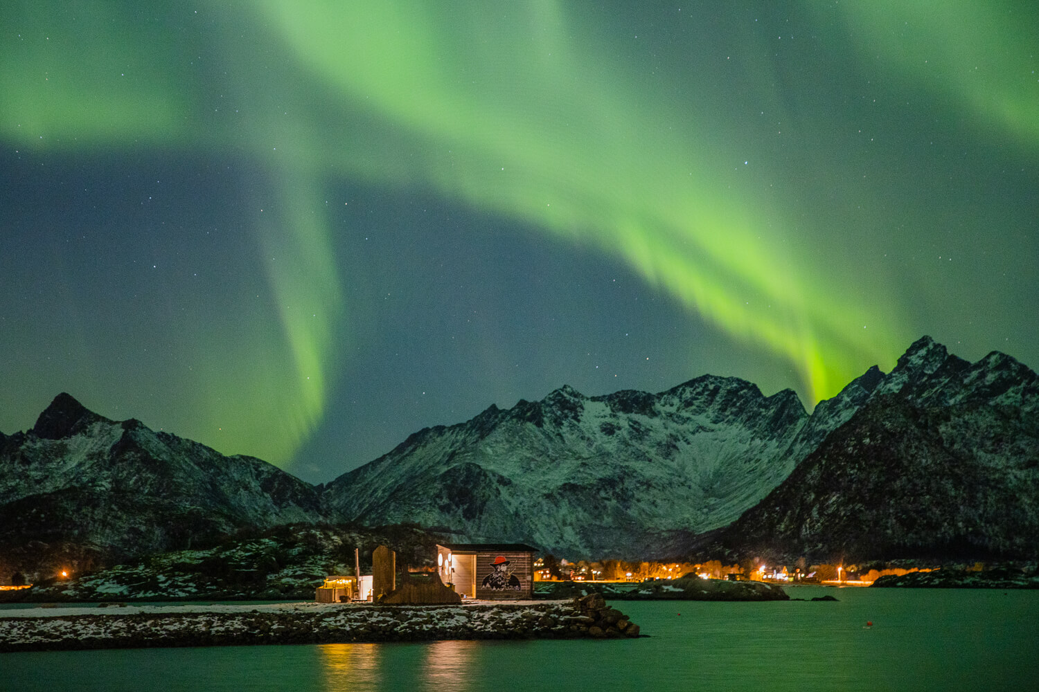 Northern lights over snowy mountains and a coastal village in Lofoten at night