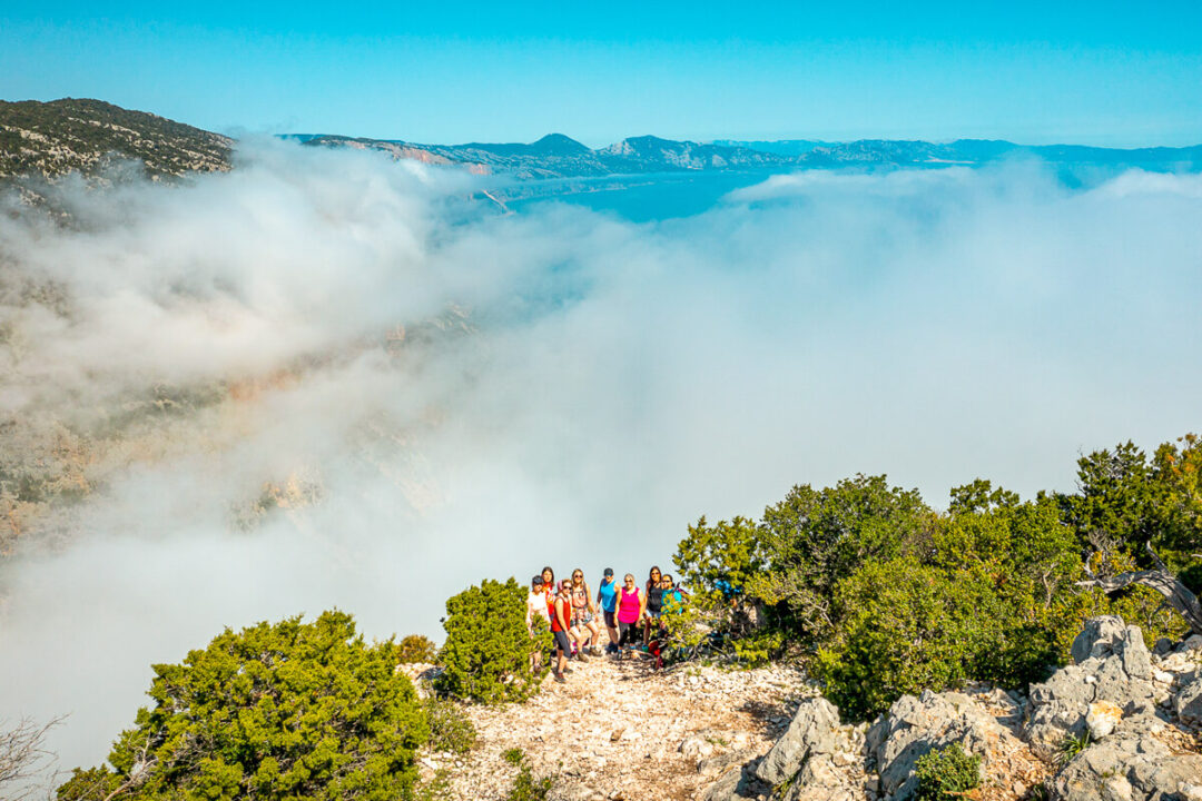 Women hiking along a coastal cliff ridge above thick sea mist and mountains.