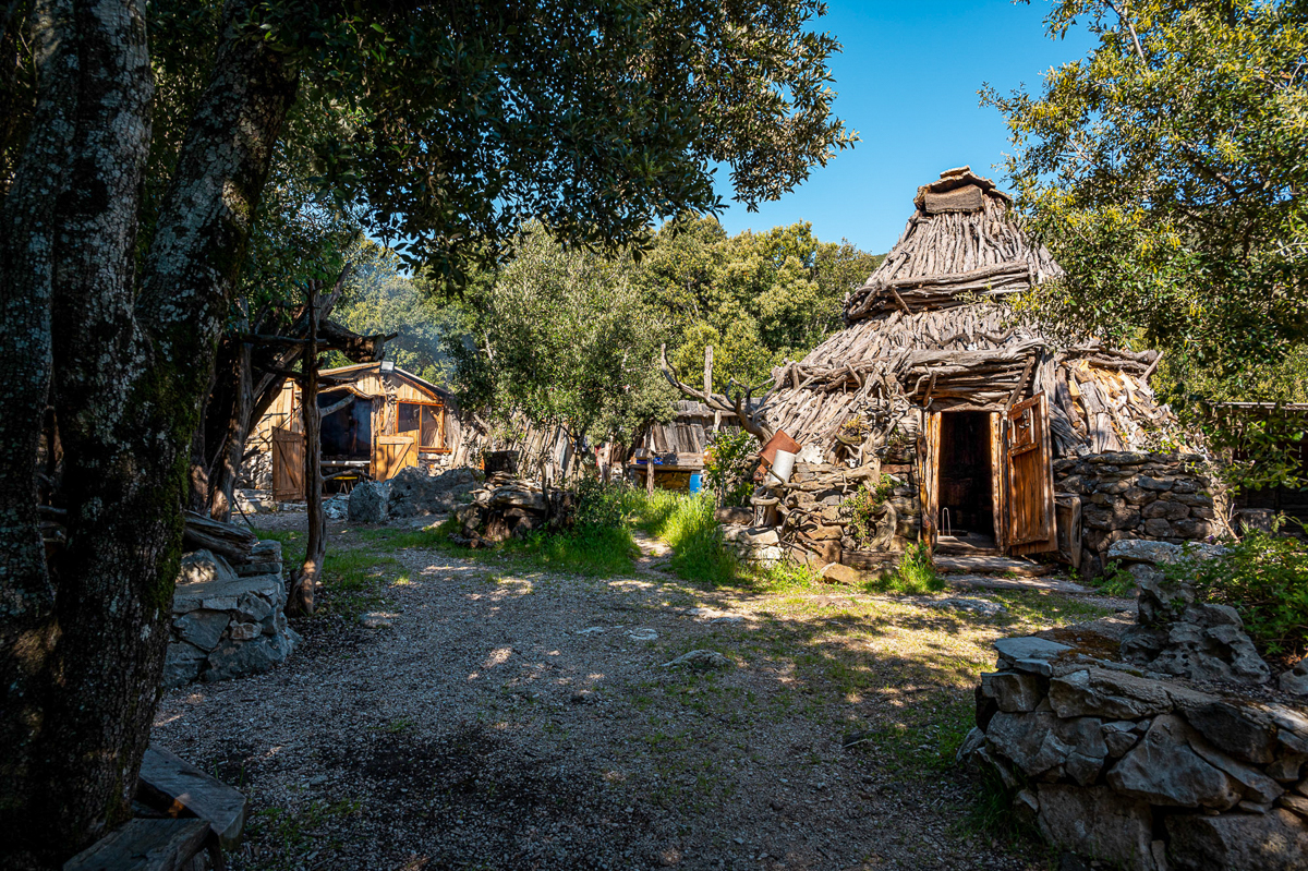 Rustic stone huts and a gravel path in a wooded village setting outdoors