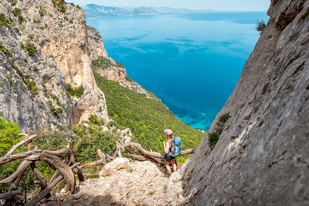 Woman hiking on rocky coastal cliffs with a backpack, overlooking turquoise sea in Sardinia
