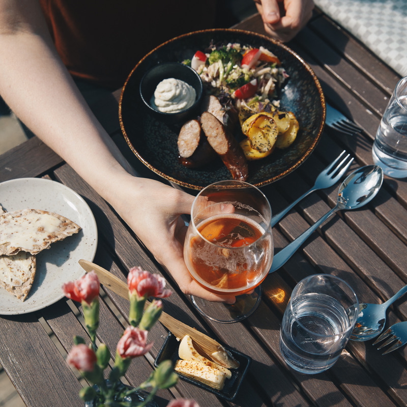 Outdoors dining table with grilled food, salad and red wine, with hands serving on a wooden bench