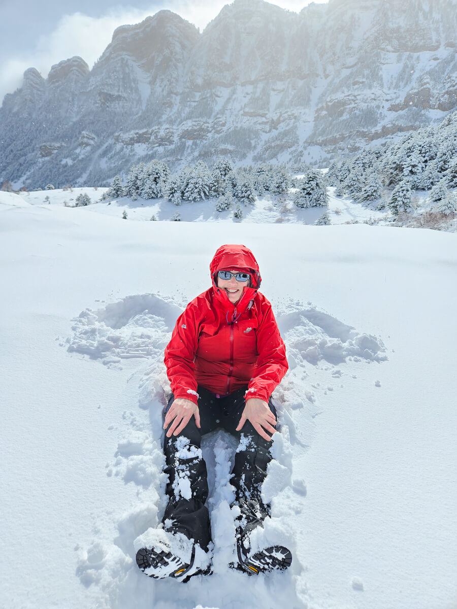 Woman in red winter gear crouching in snow with mountains behind, Pyrenees winter hiking