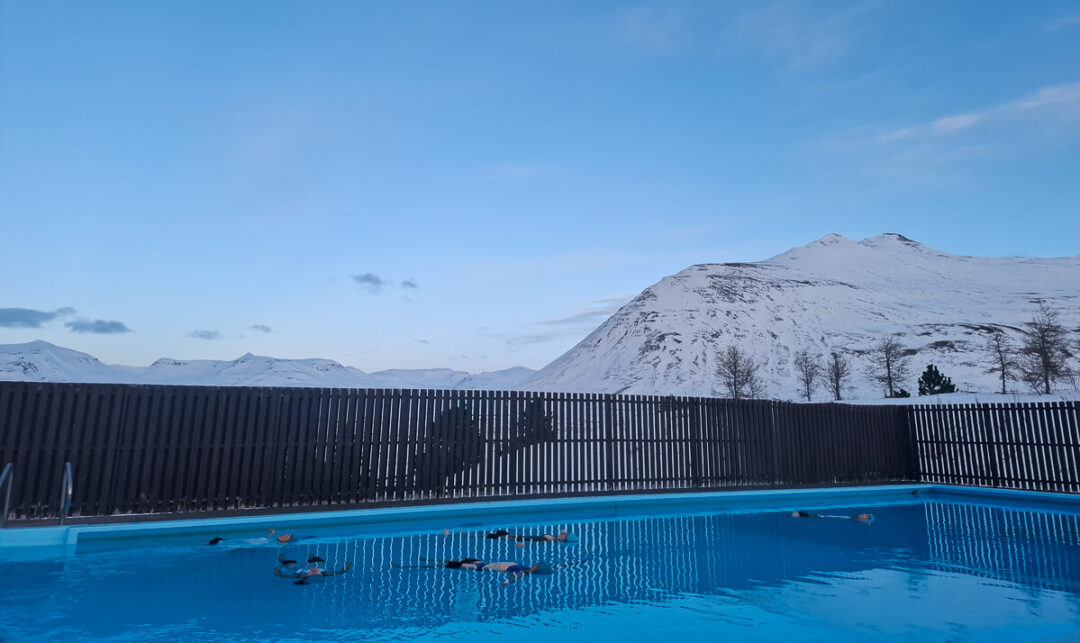 Outdoor swimming pool with snowy mountains and blue sky beyond a dark fence in winter scenery