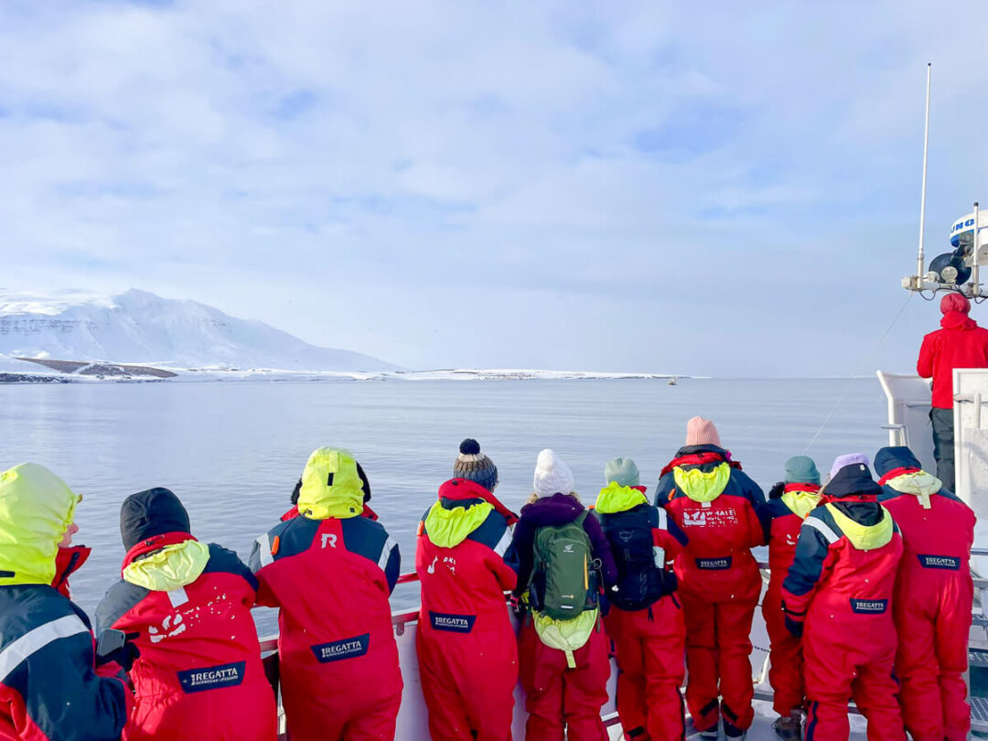 Women in red winter suits on a boat deck, facing the sea in snowy North Iceland with mountains in view