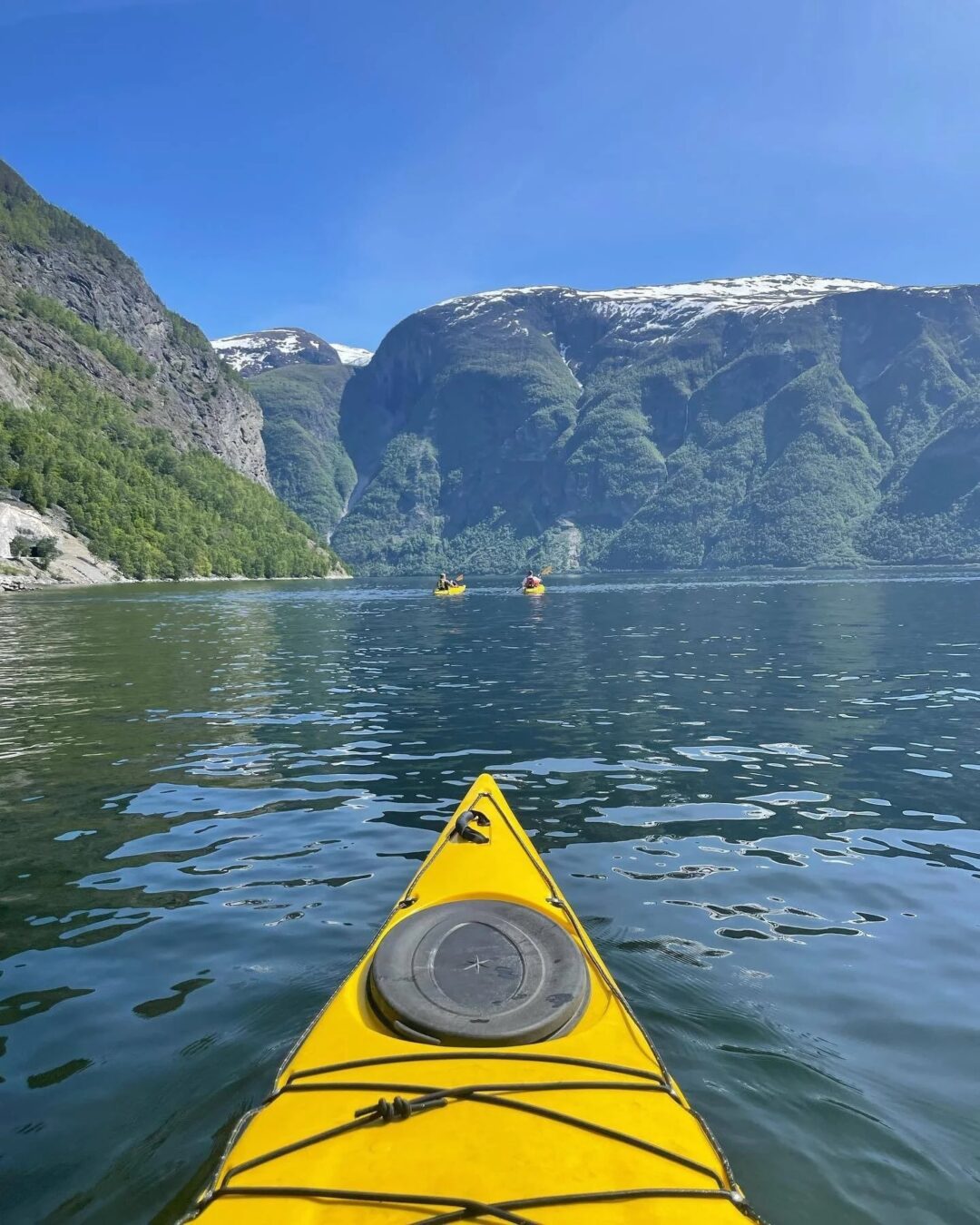 Yellow kayak bow on a calm fjord in Norway, with distant mountains and two kayaks ahead.