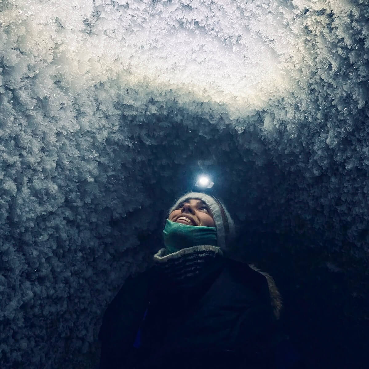 Woman in winter gear inside an icy snow cave in Svalbard, looking up in the darkness