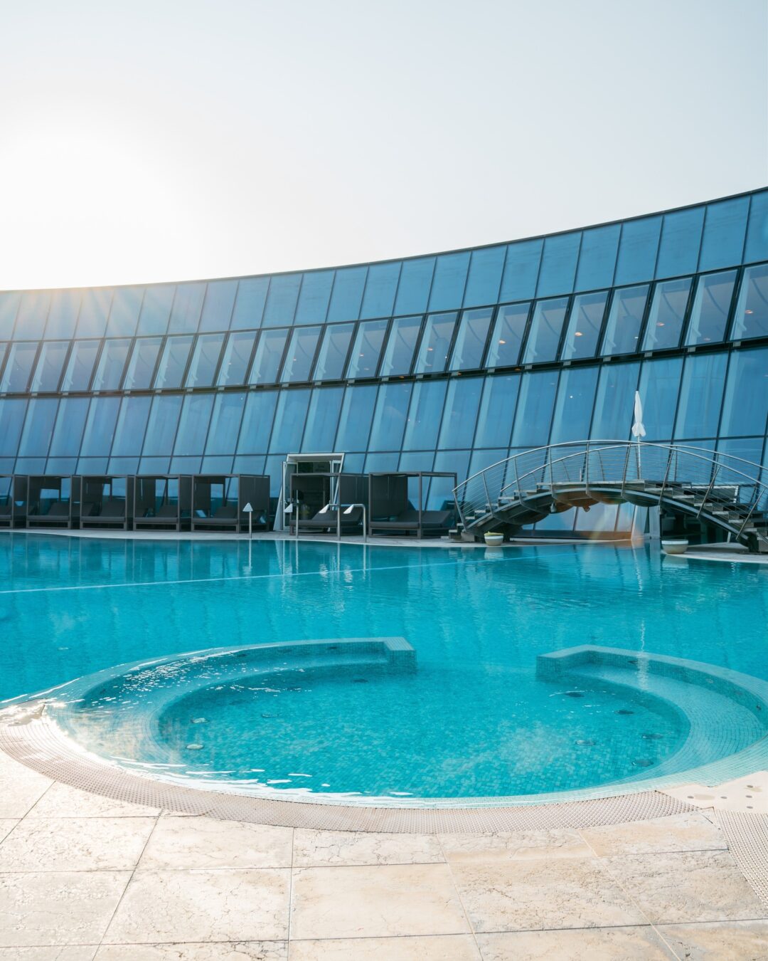 Outdoor swimming pool beside a modern glass hotel building with sun loungers and a clear blue sky