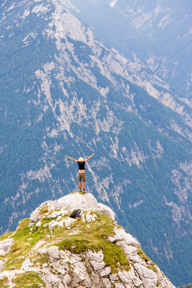 Woman hiking on a rocky summit, arms out, overlooking mountain valleys.