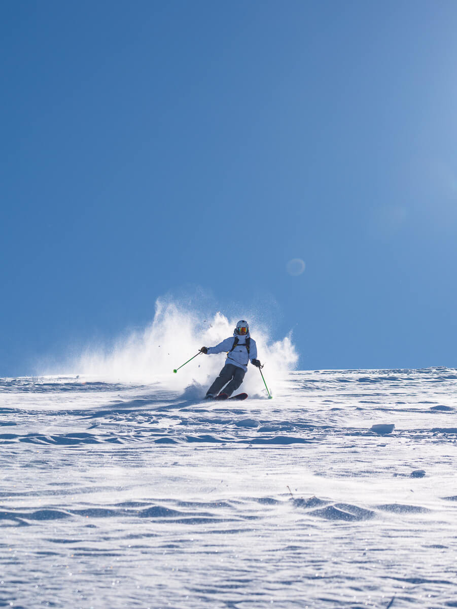 Woman ski touring through fresh snow on skis, kicking up a cloud of powder on a bright day