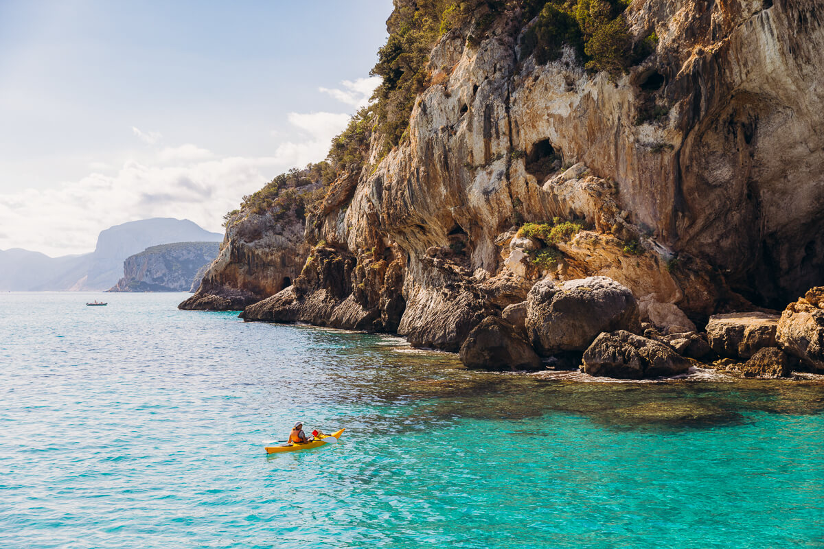 Woman kayaking in clear turquoise water beside rugged sea cliffs in East Sardinia