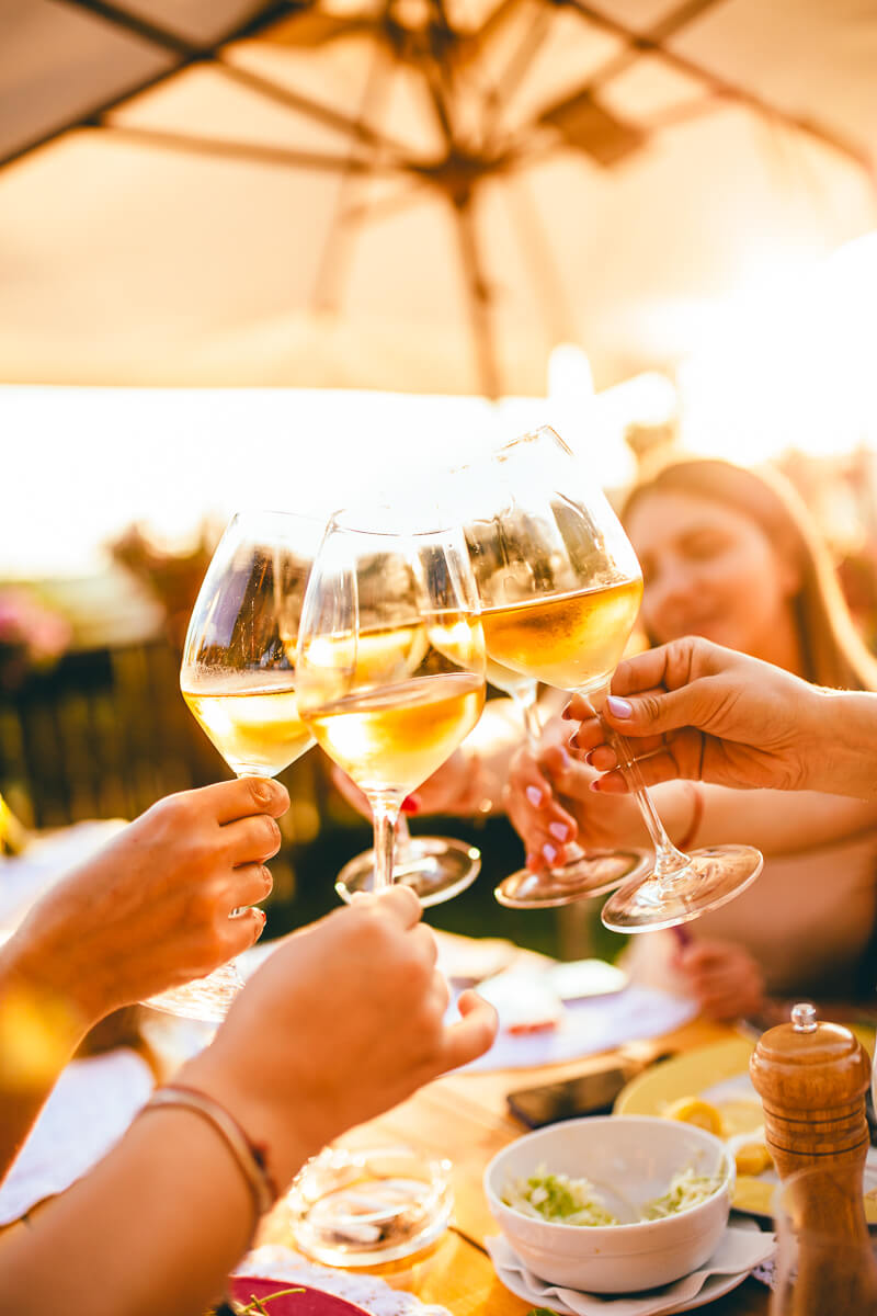 Women clinking white wine glasses at an outdoor table with umbrella, on a Lake Ohrid holiday in North Macedonia