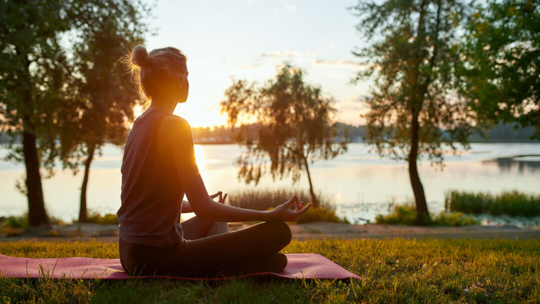 Woman meditating on a yoga mat by the lake at sunset, with trees and calm water in the background.