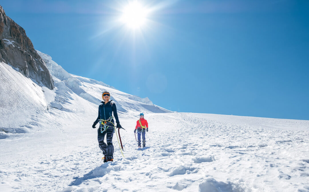 Women snowshoeing on a snowy mountain slope under a bright sun.