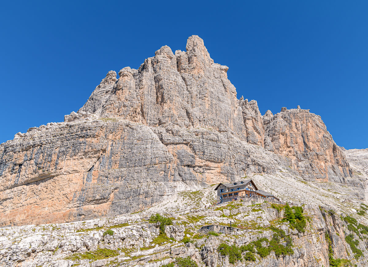 Mountain chalet in the Dolomites, Italy, beneath rugged rock peaks under a clear blue sky.