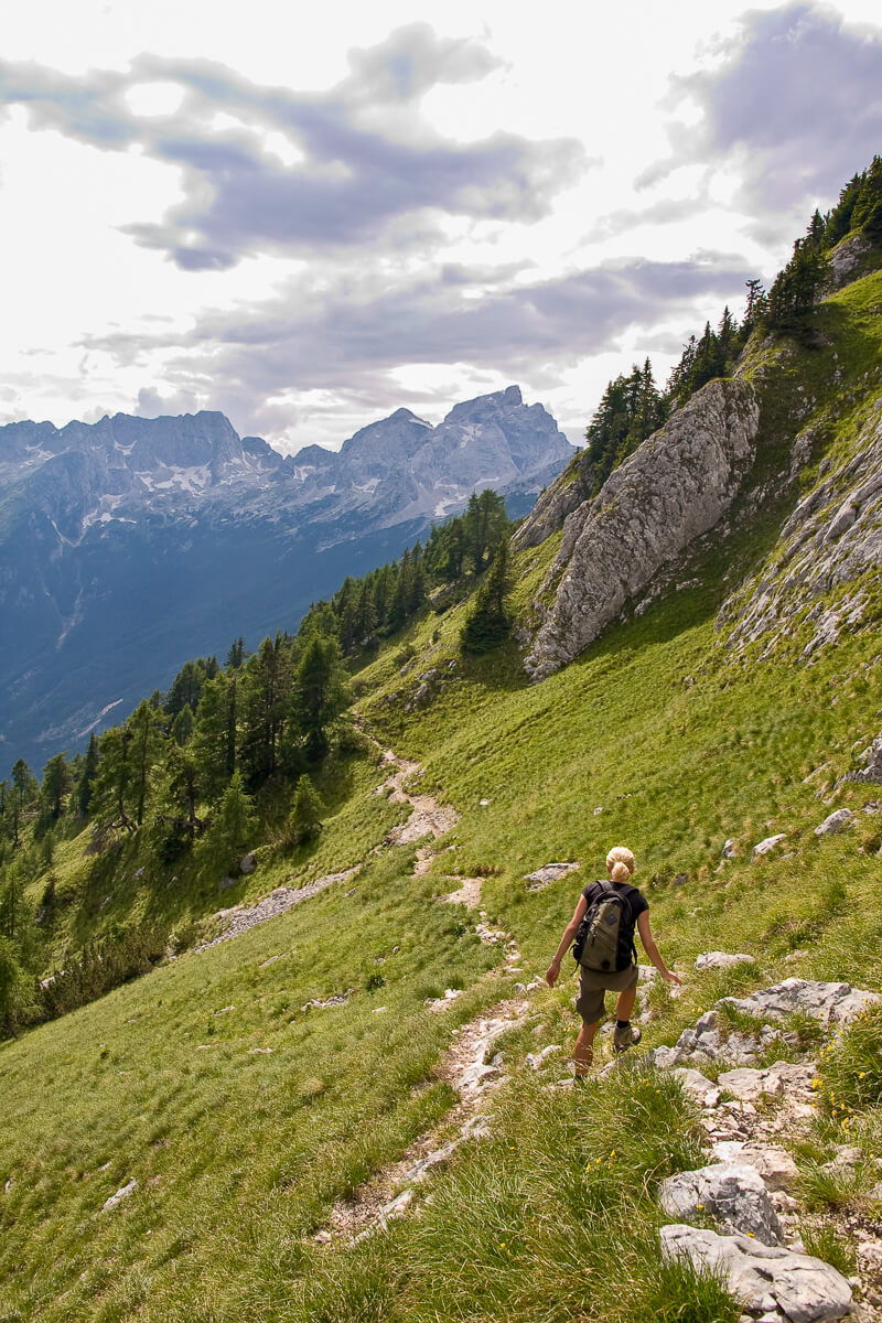 Woman hiking on an alpine mountain trail through green slopes and rocky cliffs under cloudy skies