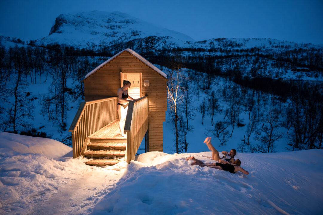 Woman standing outside a wooden cabin in snowy mountains at dusk, winter ski touring setting