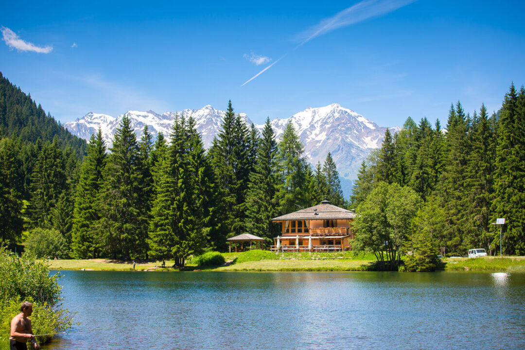 Alpine lake with a wooden chalet and pine forest, with snow-capped mountains in the background