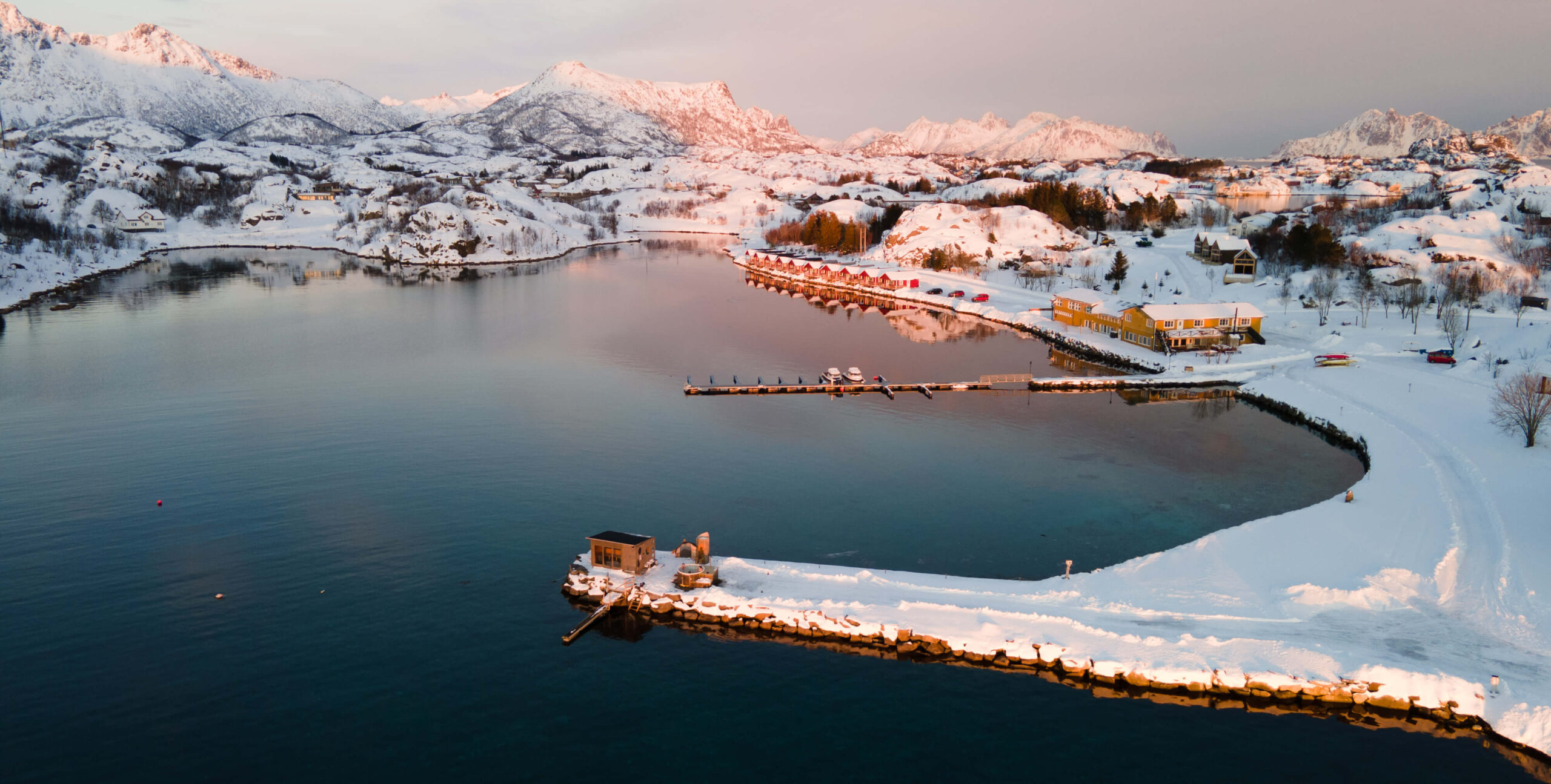 Snowy Lofoten fjord at dusk with moored boats and cabins along the shoreline