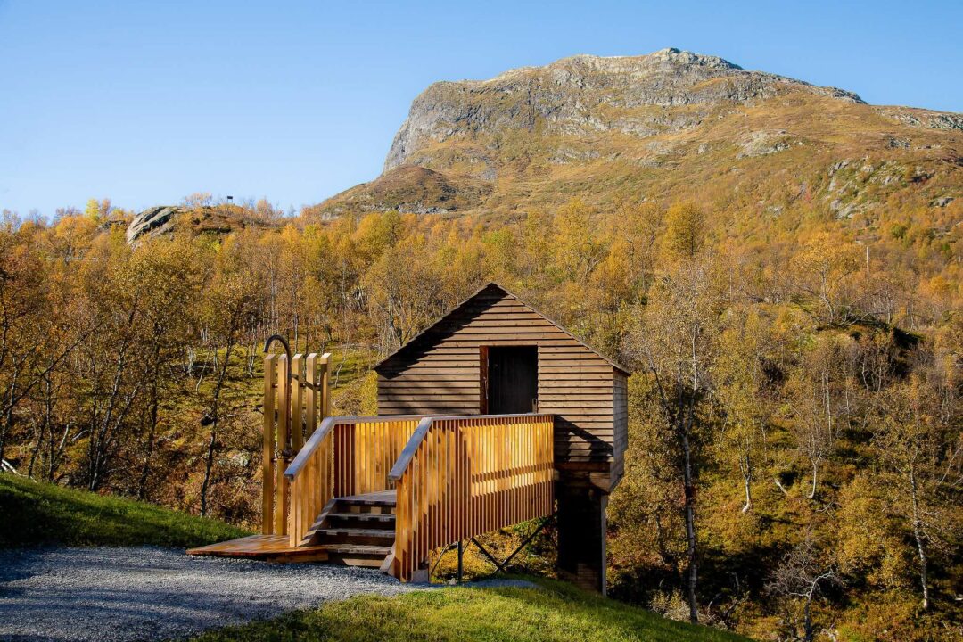 Wooden cabin beside a path in a mountainous landscape with autumn forest and a rocky peak