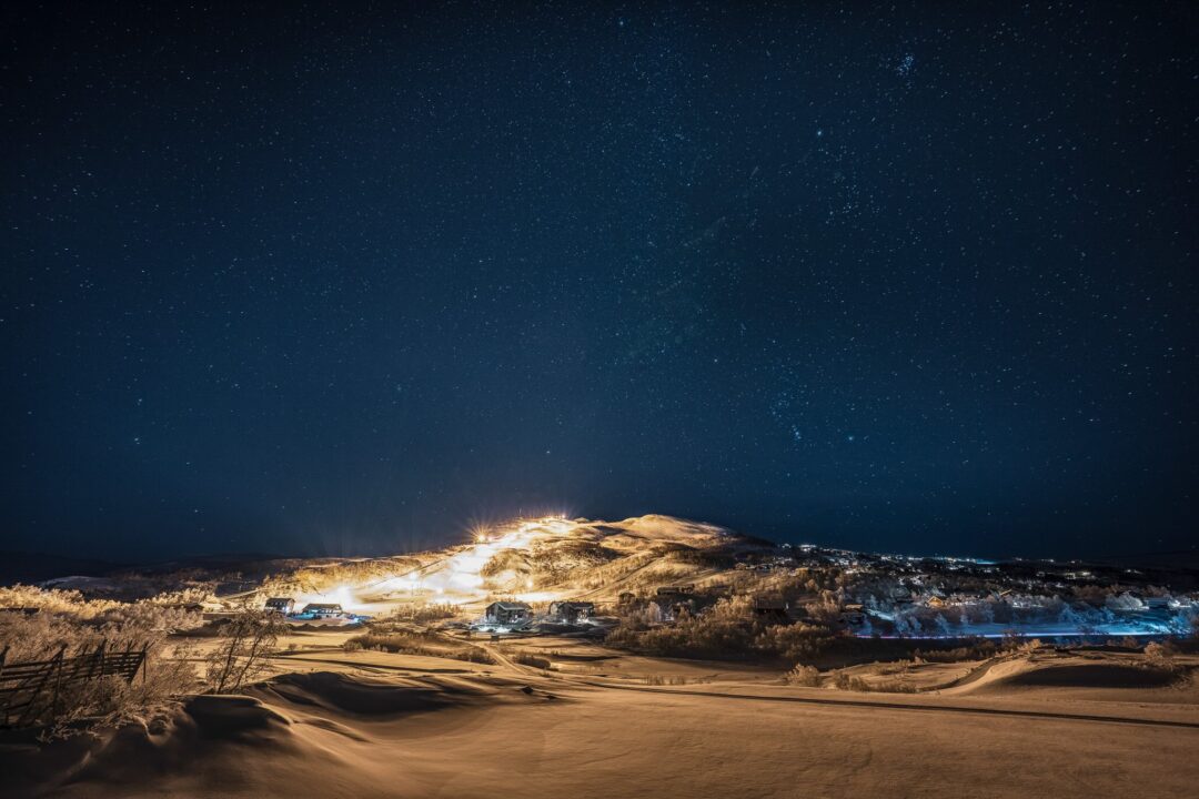 Snowy ski resort at night under a starry sky, with illuminated slopes and mountain views