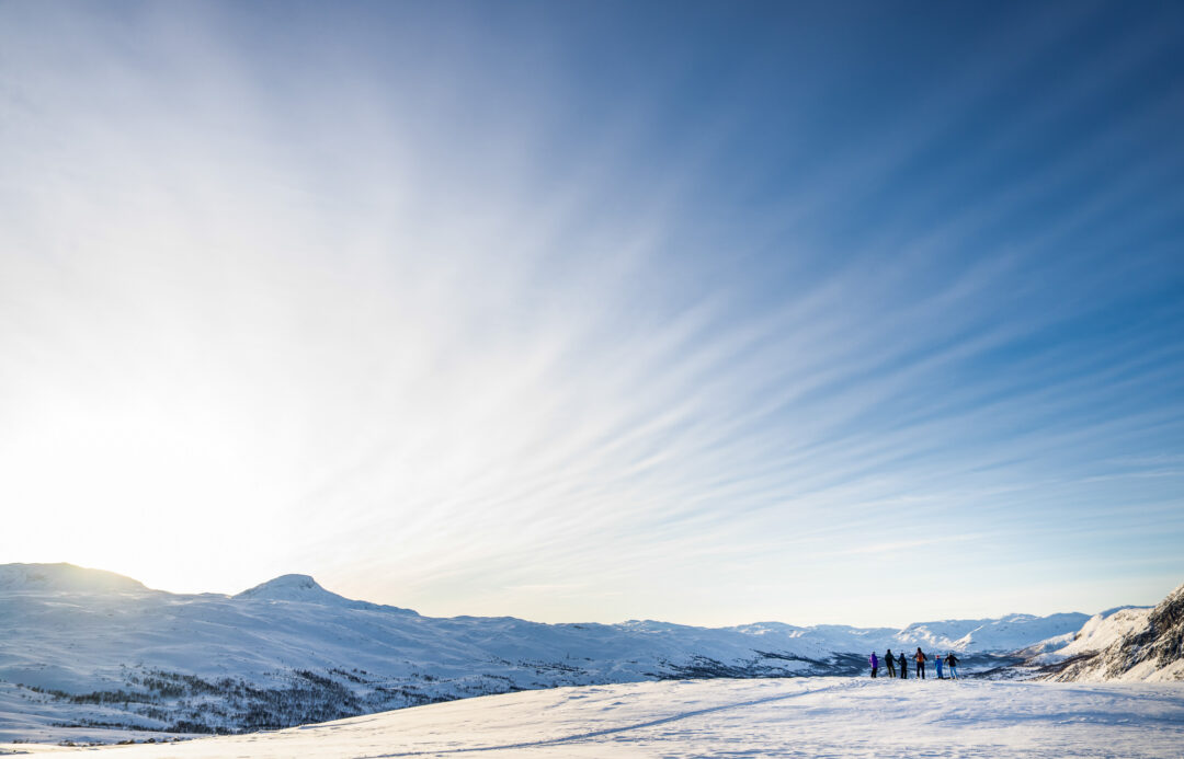 Skiers on a snowy mountain slope at dawn, with distant alpine peaks under a clear blue sky