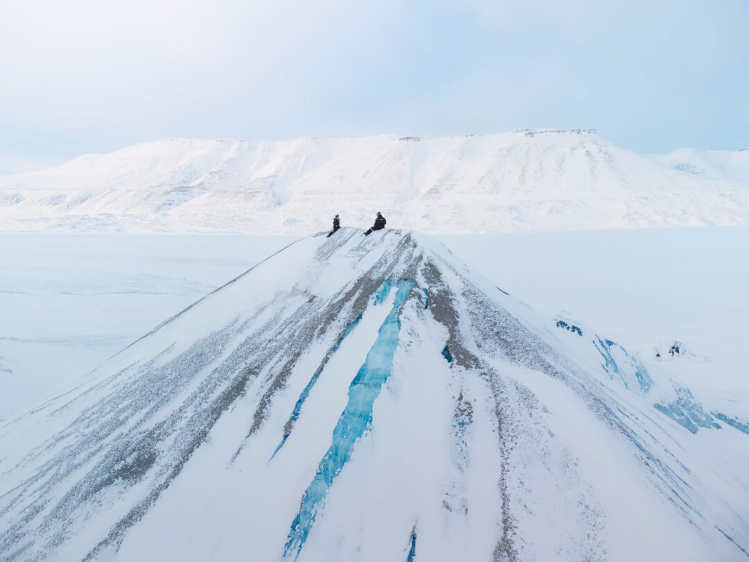 Snowmobilers ride across snow-covered mountains in Svalbard, following twin blue tracks.