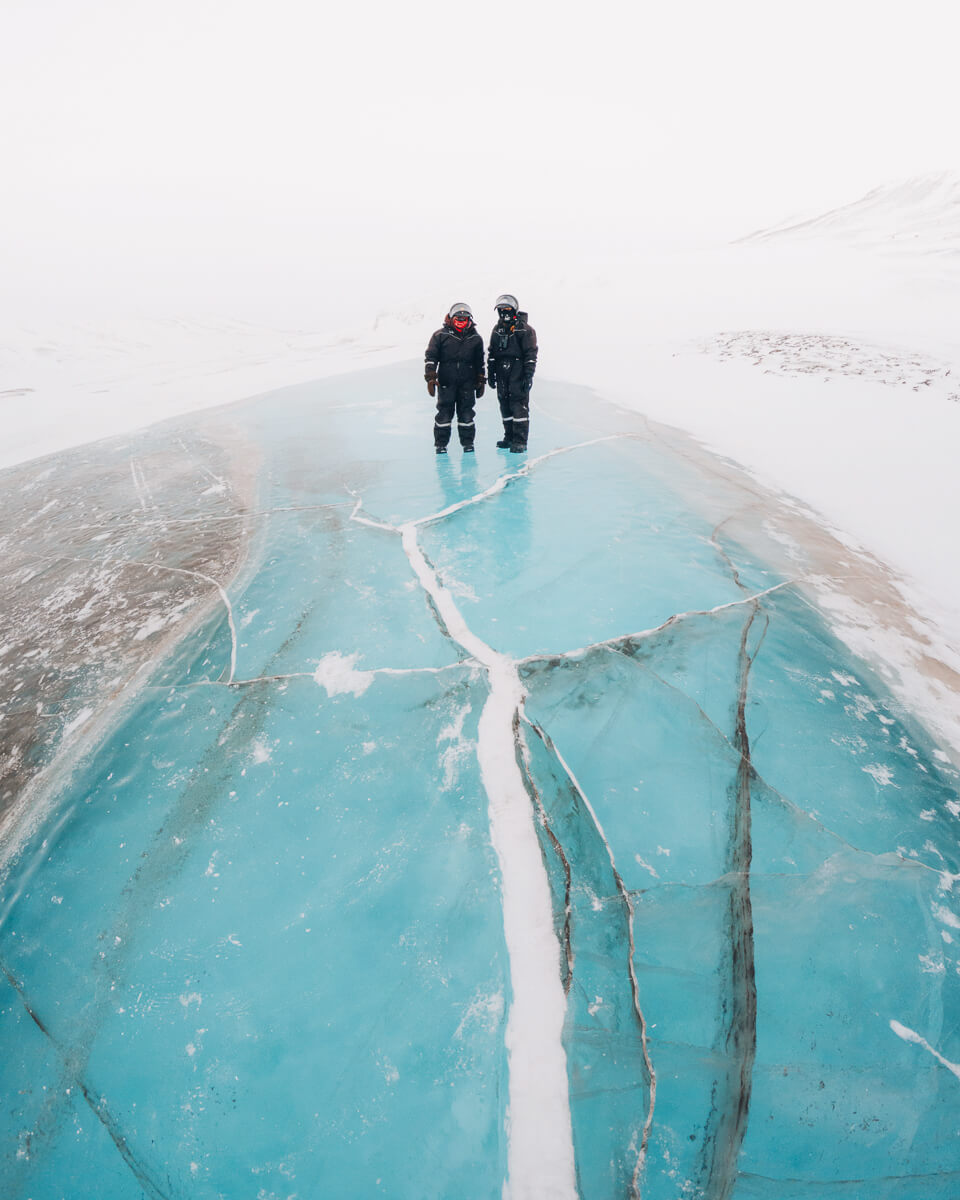 Two women in winter helmets and suits stand on blue sea ice in a snowy arctic landscape