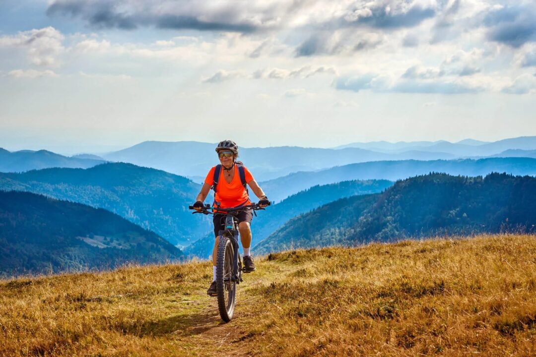 Women mountain biking on a grassy mountain trail with misty valleys and forests in the distance.