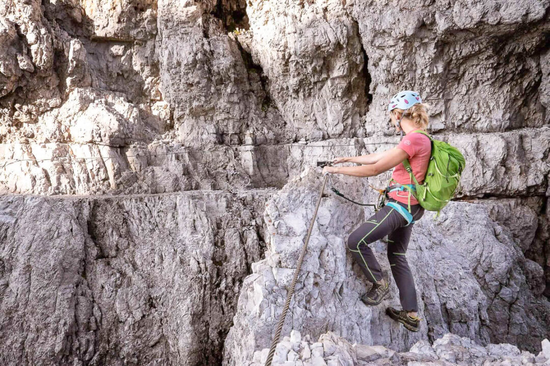 Women on a via ferrata route, climbing a rocky cliff with safety ropes and harness.