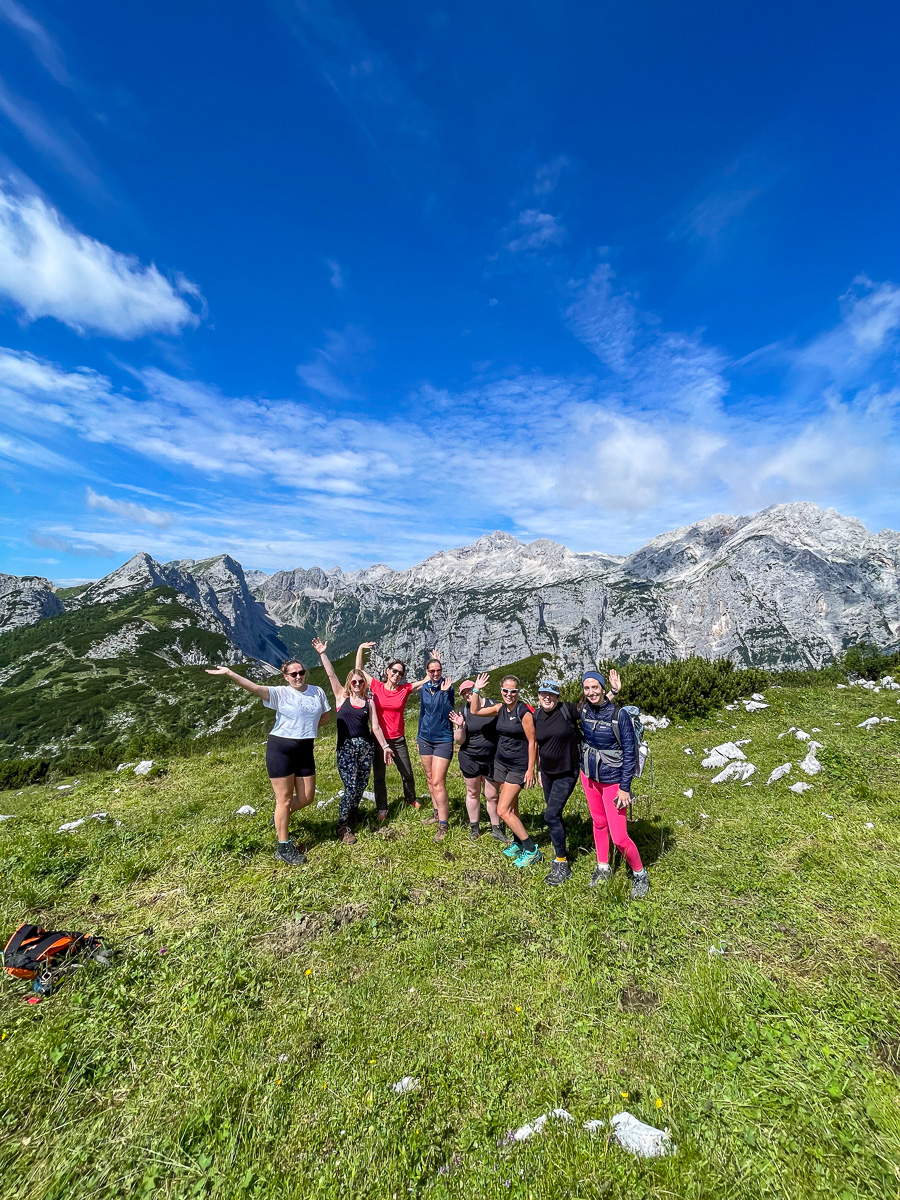 Women hiking in alpine mountains with backpacks, standing on a grassy trail under a blue sky and clouds