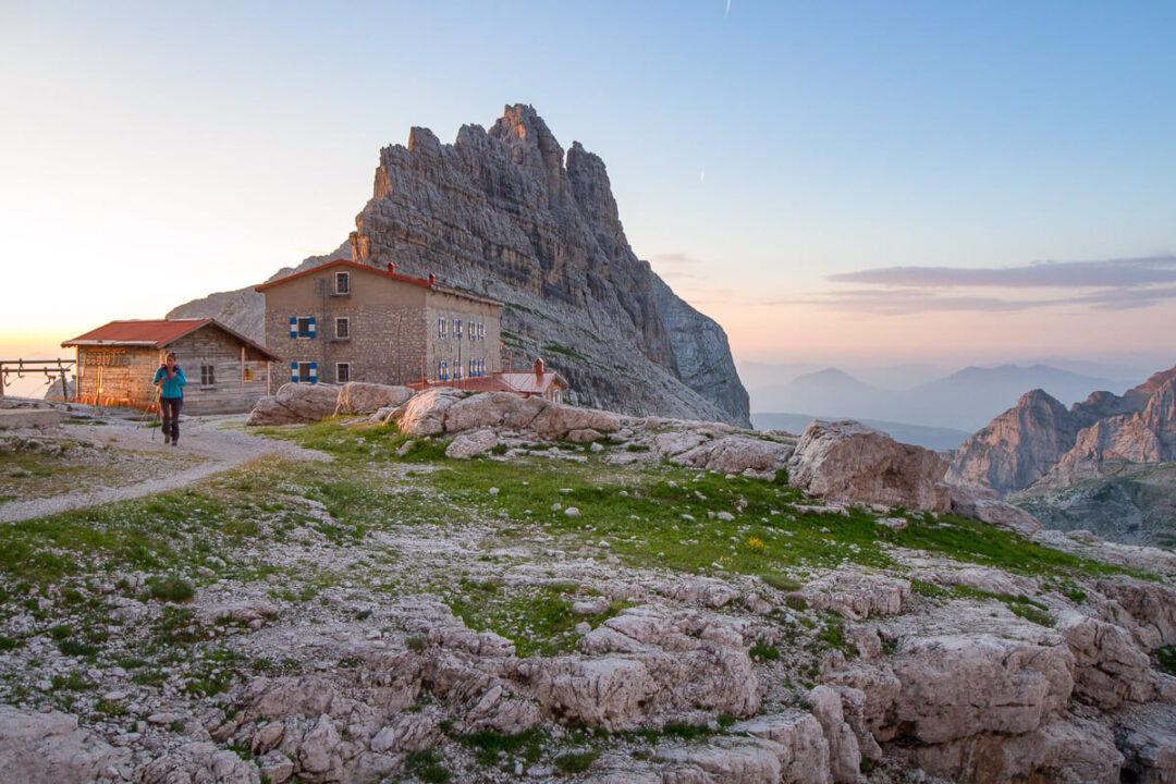 Mountain hut on a rocky ridge in the Dolomites at sunrise, with hikers on a trail.
