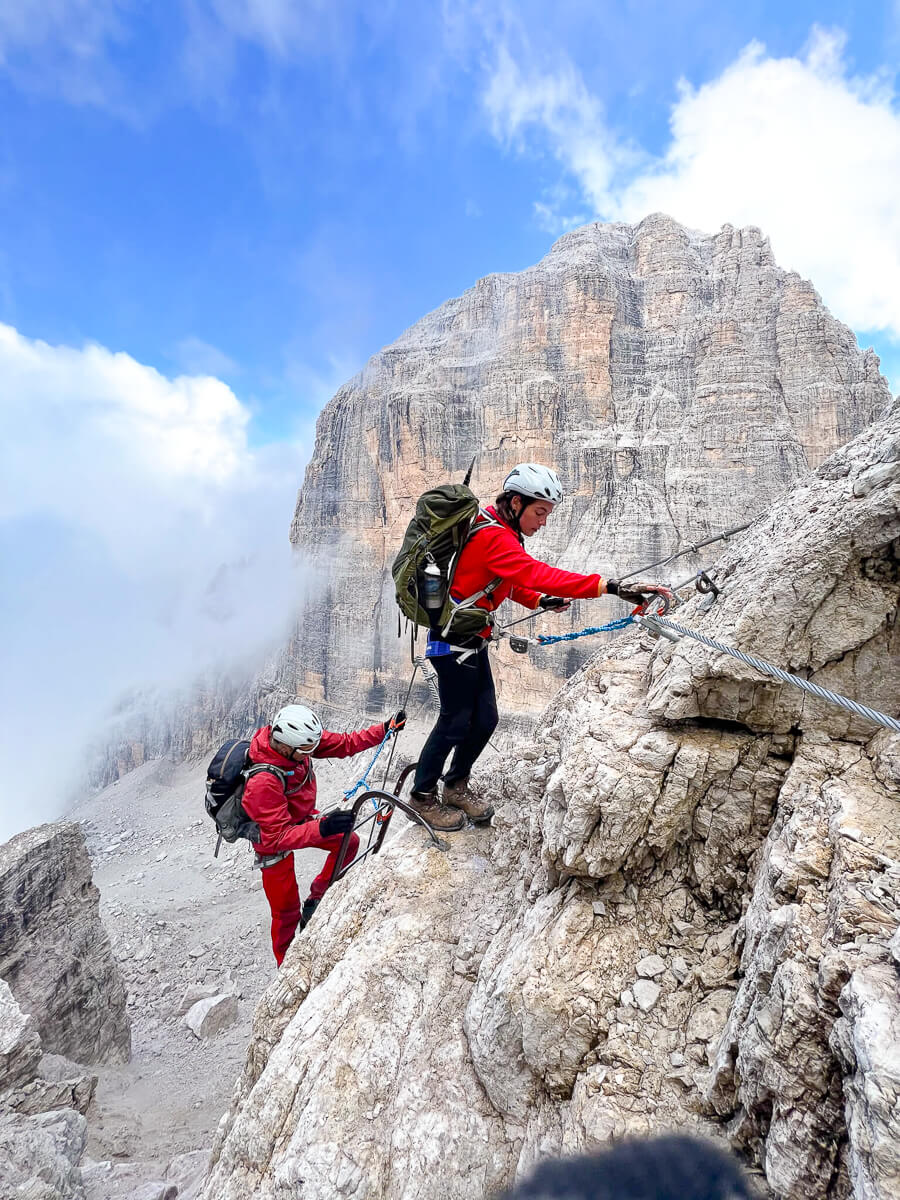 Women climbing a via ferrata along a rocky ridge in the Dolomites, with harnesses and safety cables.