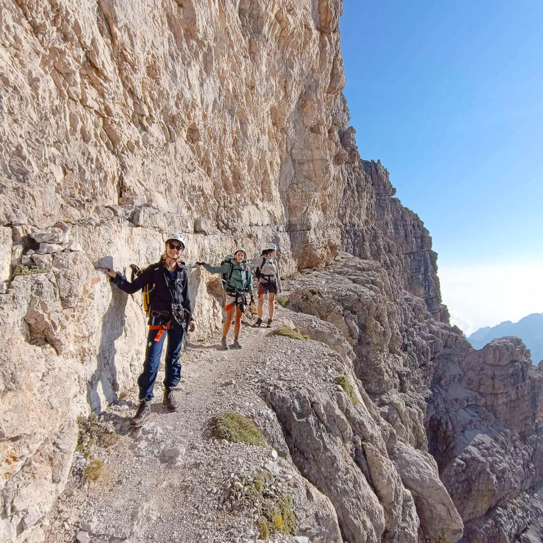 Women hiking along a via ferrata on rocky cliffs in the Dolomites, with mountain scenery in the distance.