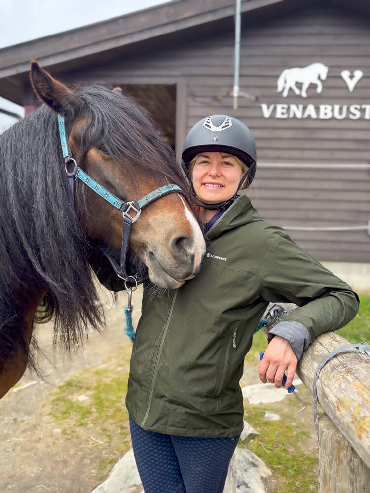 Woman horseback riding at a barn, wearing a helmet with a horse; sign reads “VENABUST”