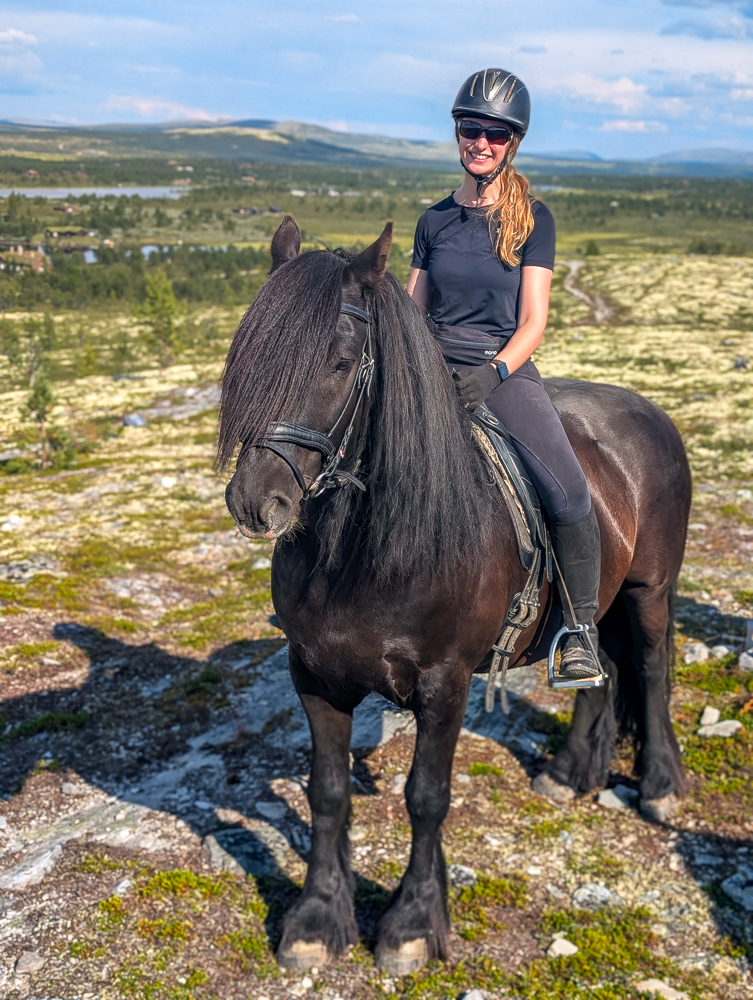 Woman on horseback outdoors, wearing a helmet and riding gear with a mountainous countryside view