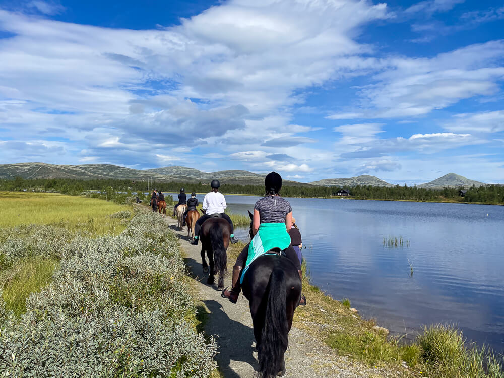 Women horseback riding along a lakeside in Norway, with mountains under a blue sky.