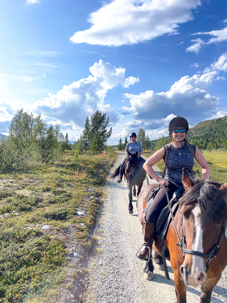Women on horseback riding along a gravel trail through forest and mountain scenery