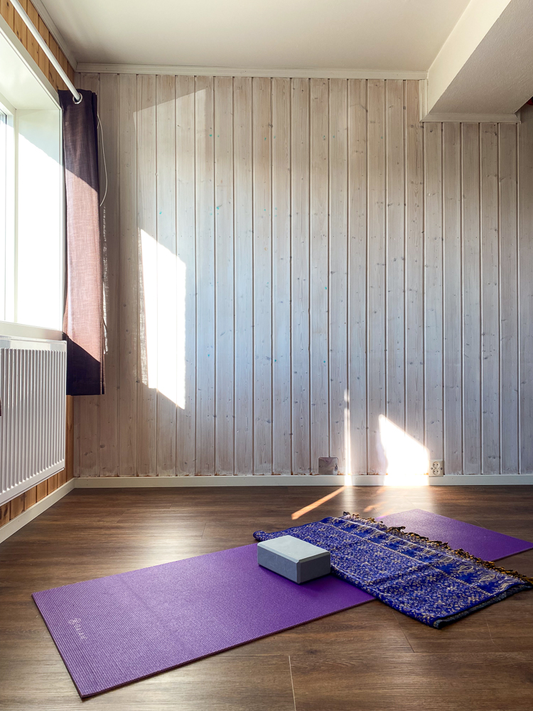 Yoga mats, blocks and a rolled blanket on a purple mat in a light-filled wooden studio.