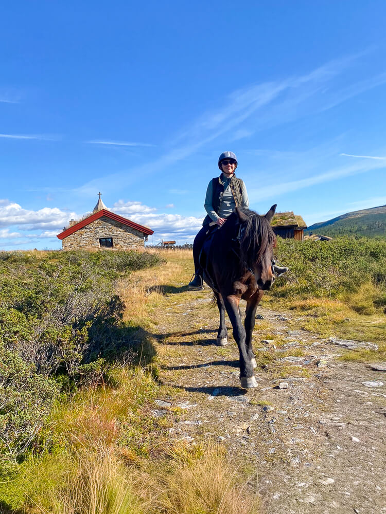 Woman horseback riding in Norway, on horseback on a trail near a small cabin