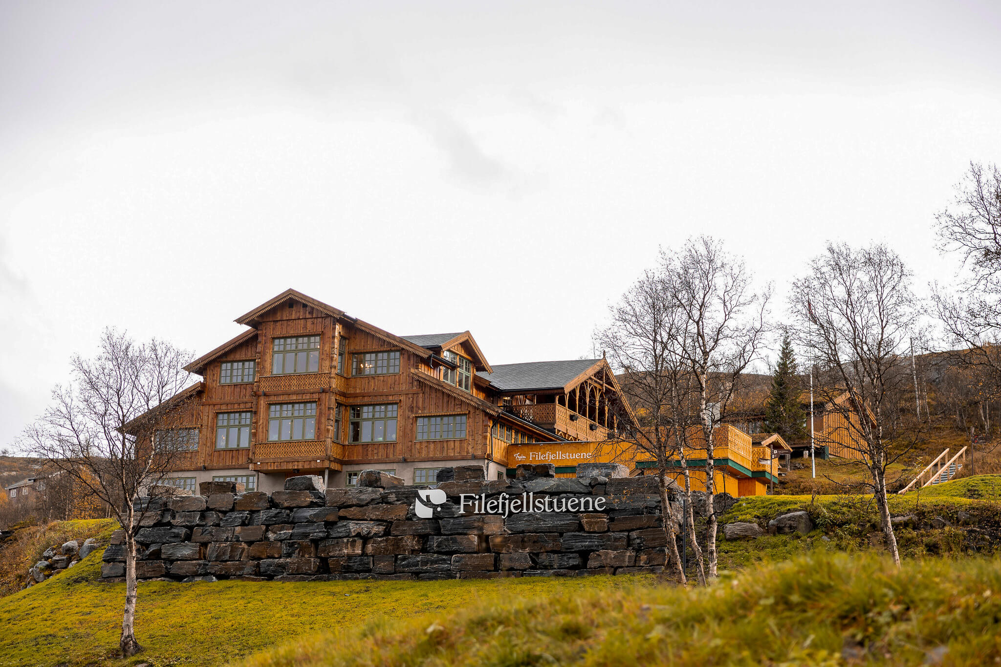 Timber mountain lodge with stone wall and sign “Fjellestuene” with grassy countryside in the background