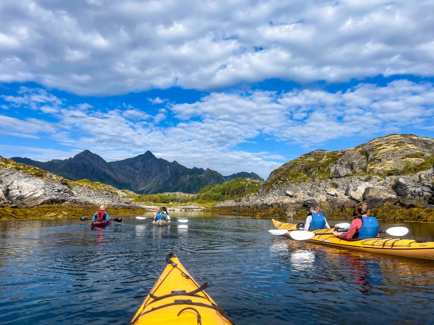Women kayaking on calm water with paddles, surrounded by rocky fjord mountains under a cloudy sky