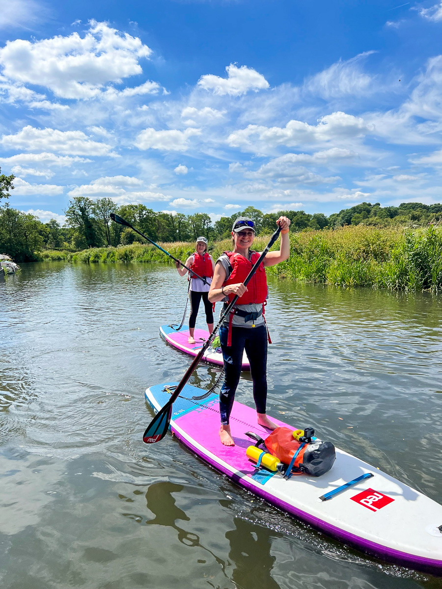 Women beginner paddleboarding on a calm river, standing on pink stand-up paddleboards under blue sky