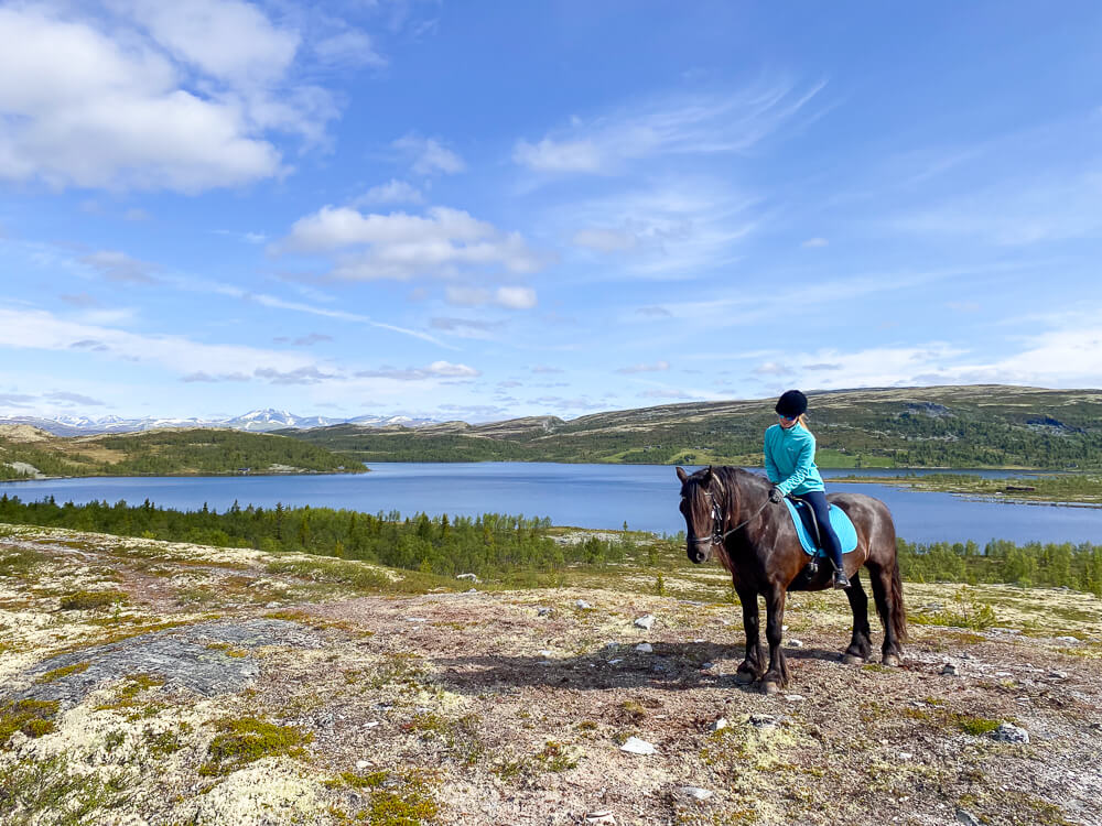 Woman horseback riding on a hillside overlooking a Norwegian lake and mountains.